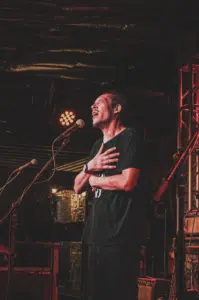 A man stands on stage with his hands on his chest, singing passionately into a microphone under stage lighting at one of the vibrant festivals.